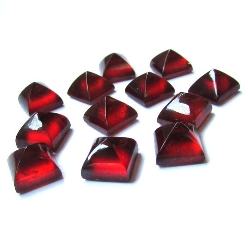 May include: A collection of faceted, square-shaped gemstones in a deep red color. The gems have a glossy finish and are arranged on a white surface, with light reflecting off their surfaces. These could be used for jewelry making or other crafts.