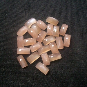 Peut inclure: Une collection de 18 pierres de lune rectangulaires lisses de couleur rose pâle. Les pierres ont un léger chatoiement et sont disposées en tas sur un fond sombre.