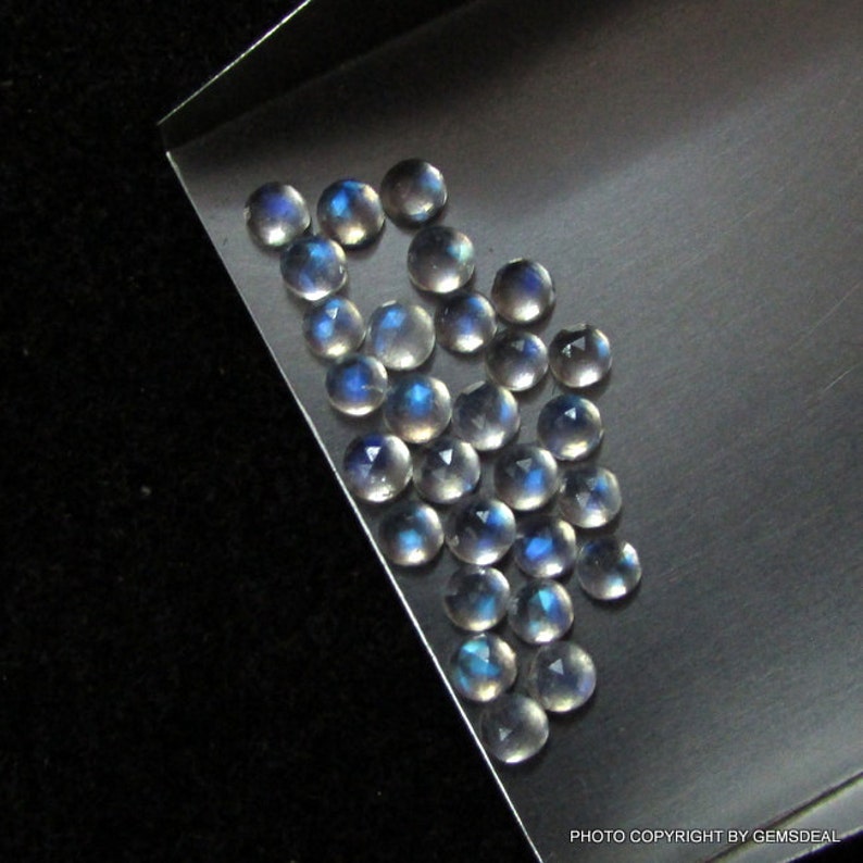 Puede incluir: Una colecci&oacute;n de piedras preciosas de piedra lunar redondas e iridiscentes de varios tama&ntilde;os. Las piedras son de color blanco azulado p&aacute;lido con un brillo brillante y opalescente.