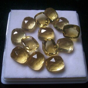 May include: A collection of 12 faceted, golden yellow gemstones displayed in a white, rectangular box. The gemstones are all cut in a similar, cushion-shaped style.