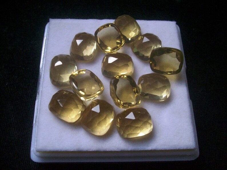 May include: A collection of 12 faceted, golden yellow gemstones displayed in a white, rectangular box. The gemstones are all cut in a similar, cushion-shaped style.