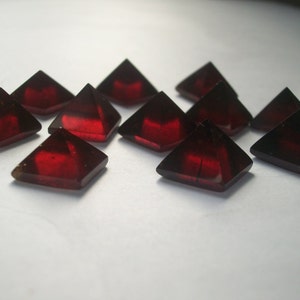 May include: A group of ten red gemstone pyramids, each with a four-sided base and a pointed top. The pyramids are arranged in a loose cluster on a white surface.