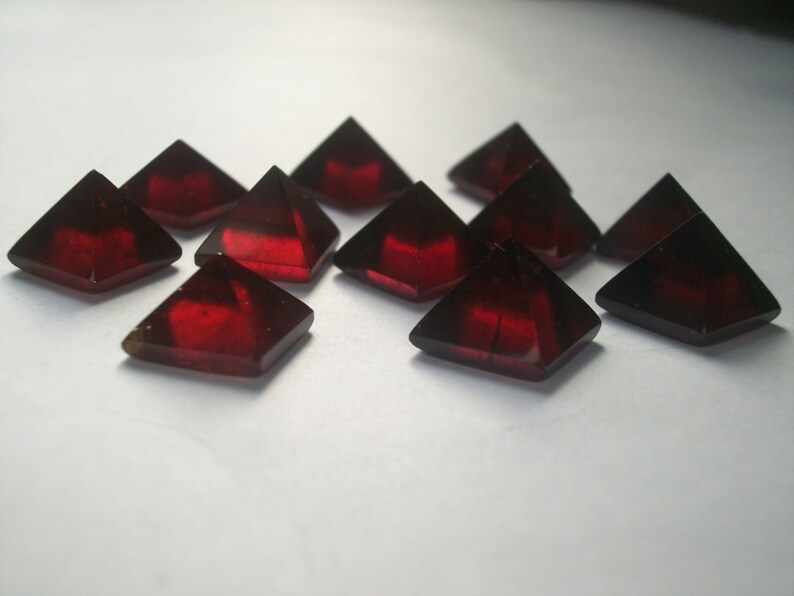 May include: A group of ten red gemstone pyramids, each with a four-sided base and a pointed top. The pyramids are arranged in a loose cluster on a white surface.