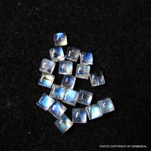 K&ouml;nnte beinhalten: Eine Sammlung von 15 regenbogenfarbenen blauen Mondstein-Cabochons, die jeweils in quadratischer Form geschnitten sind. Die Steine liegen lose auf einem schwarzen Hintergrund.