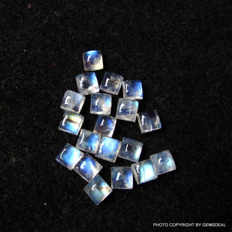 K&ouml;nnte beinhalten: Eine Sammlung von 15 regenbogenfarbenen blauen Mondstein-Cabochons, die jeweils in quadratischer Form geschnitten sind. Die Steine liegen lose auf einem schwarzen Hintergrund.
