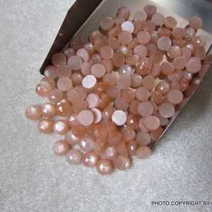 May include: A collection of approximately 50 peach-colored sunstone cabochons in various sizes and shapes. The stones are smooth and have a shimmery, iridescent quality.