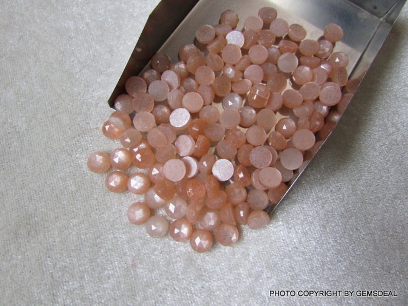 May include: A collection of approximately 50 peach-colored sunstone cabochons in various sizes and shapes. The stones are smooth and have a shimmery, iridescent quality.