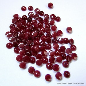 May include: A scattering of small, polished, deep red gemstones. The round stones are spread across a white surface, with light reflecting off their smooth surfaces. The image is a close-up, highlighting the details of the gemstones.