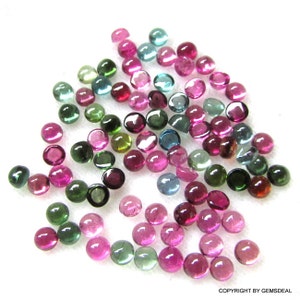 May include: A collection of round, faceted gemstones in various shades of green, pink, and blue. The gemstones are scattered on a white surface.