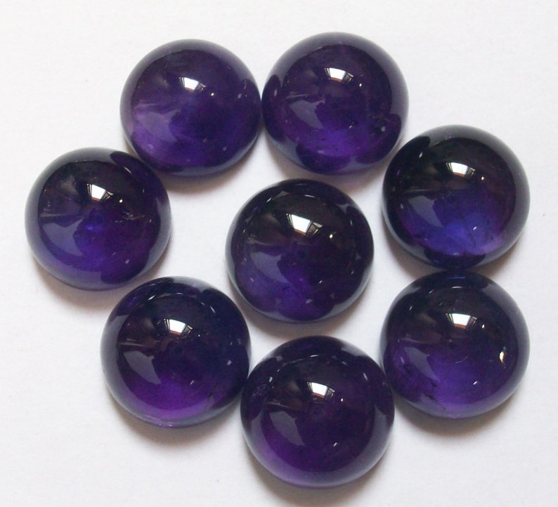 May include: Eight round, polished amethyst cabochons in various shades of purple. The cabochons are arranged in a circular pattern on a white surface.