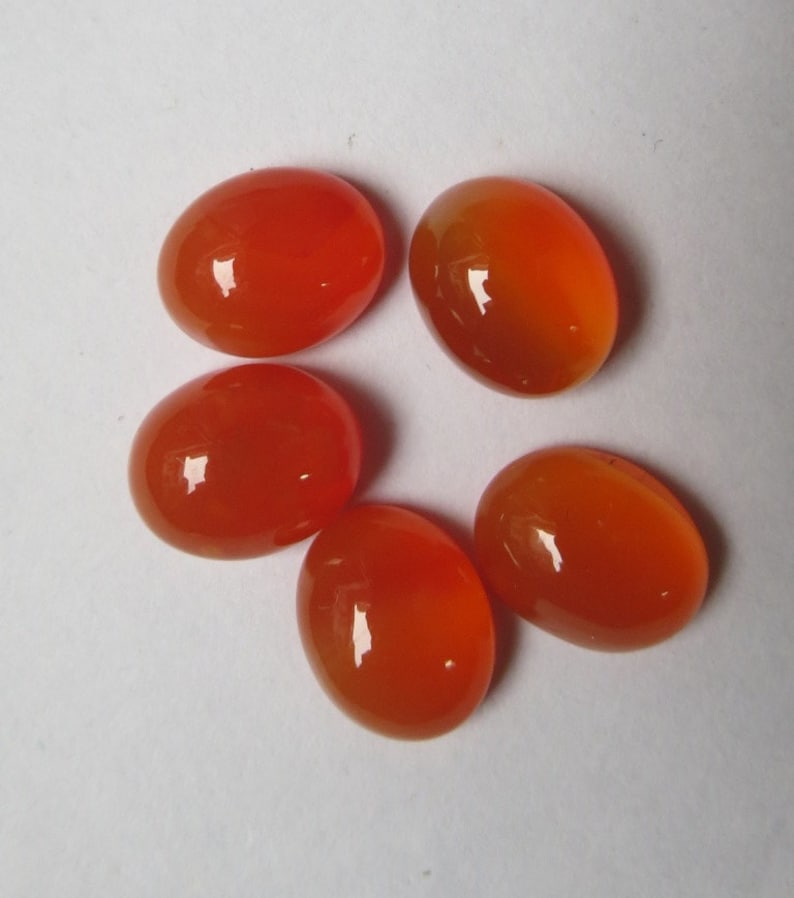 May include: Five oval-shaped carnelian gemstones, each with a smooth, polished surface and a vibrant orange color.