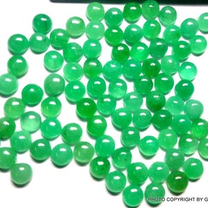 May include: A collection of round, green emerald gemstones. The gemstones are smooth and polished, and they are scattered on a white surface.