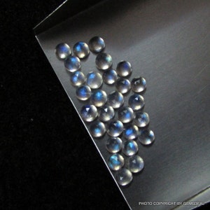 Puede incluir: Una colecci&oacute;n de 20 cabujones de piedra lunar iridiscente, cada uno con un brillo azul &uacute;nico. Las piedras son redondas y tienen una superficie lisa y pulida. Est&aacute;n dispuestas en una pila suelta sobre una bandeja plateada.