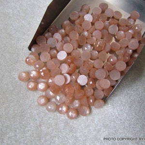 May include: A pile of round, peach-colored sunstone cabochons with a shimmery, iridescent finish. The stones are loose and scattered on a silver tray.
