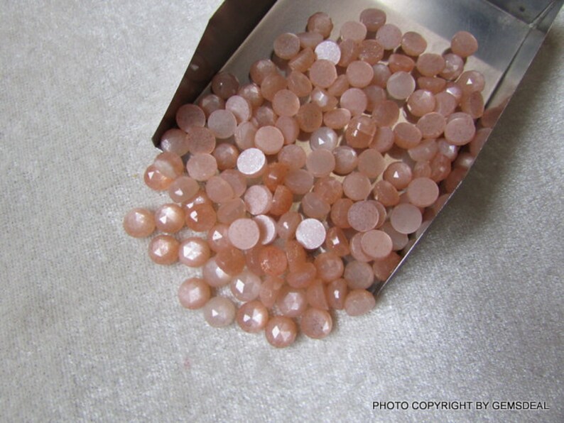 May include: A pile of round, peach-colored sunstone cabochons with a shimmery, iridescent finish. The stones are loose and scattered on a silver tray.