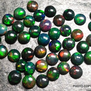 May include: A collection of 30 round black opals with green, red, and blue iridescent flashes. The opals are arranged in a random pattern on a gray surface.