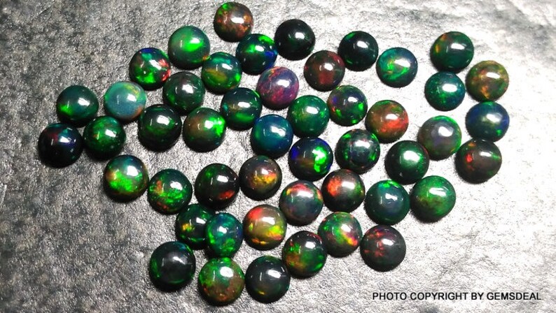 May include: A collection of 30 round black opals with green, red, and blue iridescent flashes. The opals are arranged in a random pattern on a gray surface.