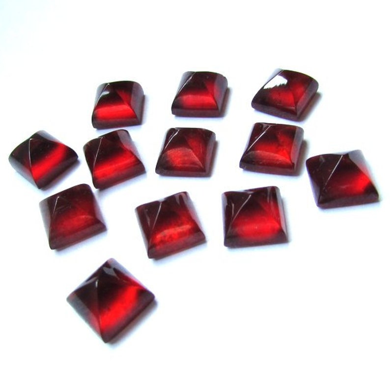 May include: A collection of eleven square, faceted, dark red gemstones. Each gem has a pyramidal shape with a lighter red center, creating a striking contrast. The gems are scattered on a white background, showcasing their color and form.