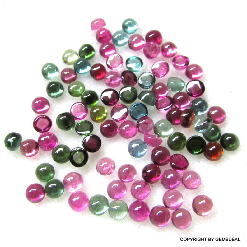 May include: A collection of round, faceted, multi-colored gemstones. The gemstones are in shades of pink, green, blue, and brown. The gemstones are arranged in a random pattern on a white surface.