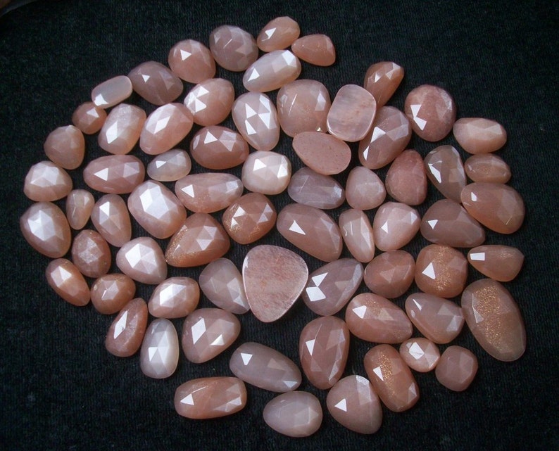 May include: A collection of approximately 50 loose, faceted, peach-colored sunstone gemstones. The stones are various sizes and shapes, with a pearlescent sheen.