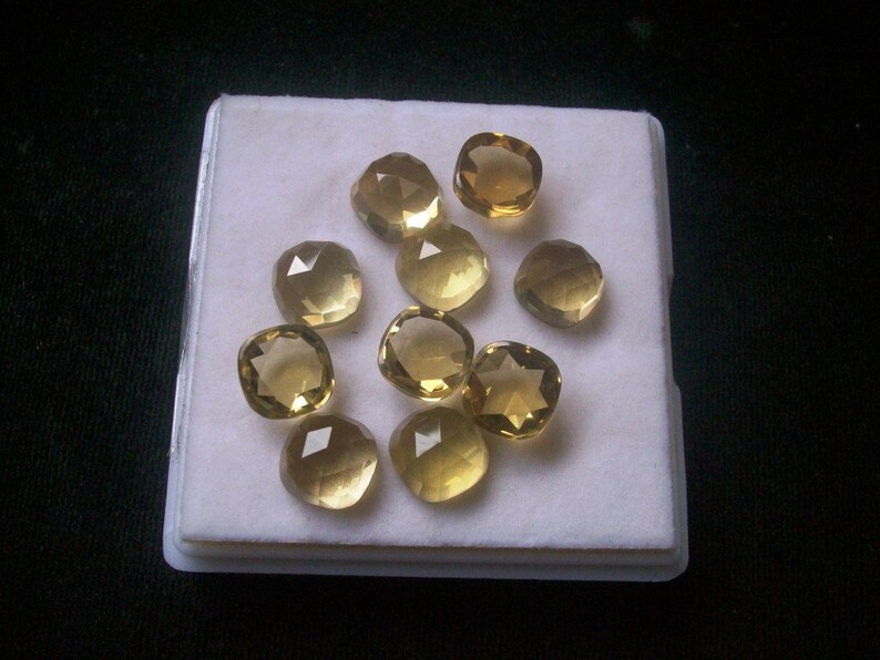 May include: A collection of ten faceted, yellow-gold gemstones displayed on a white background. The gemstones are all approximately the same size and shape, and they have a smooth, polished finish.
