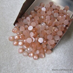 May include: A pile of small, round, peach-colored sunstone cabochons in a metal tray. The stones have a shimmery, iridescent finish.