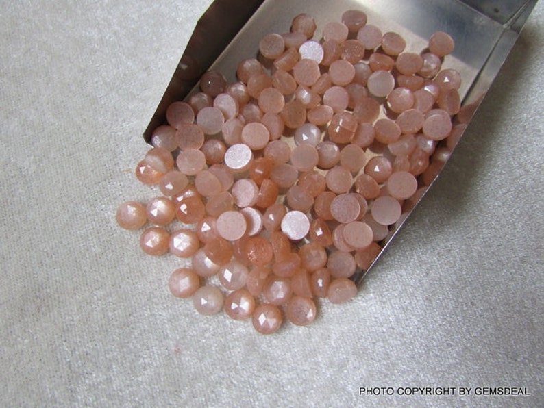 May include: A pile of small, round, peach-colored sunstone cabochons in a metal tray. The stones have a shimmery, iridescent finish.