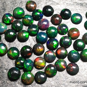 May include: A collection of round, black opal cabochons with green, red, and blue iridescent flashes. The opals are arranged in a random pattern on a gray surface.
