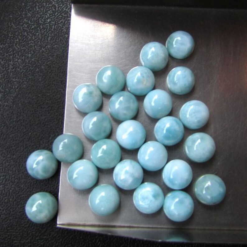 May include: A collection of 20 round, light blue cabochon gemstones. The gemstones are arranged in a triangular shape on a silver surface.