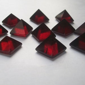 May include: A group of twelve red, faceted, glass pyramid-shaped crystals. The crystals are arranged in a random pattern on a white surface.