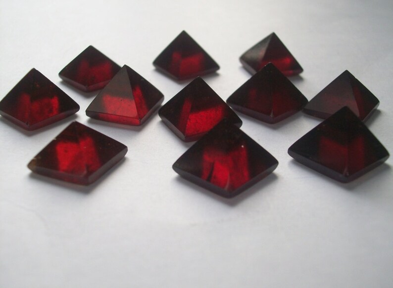 May include: A group of twelve red, faceted, glass pyramid-shaped crystals. The crystals are arranged in a random pattern on a white surface.