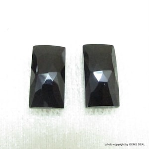 Peut inclure: Deux pierres précieuses rectangulaires noires avec une taille facetée. Les pierres précieuses sont sur une surface blanche.