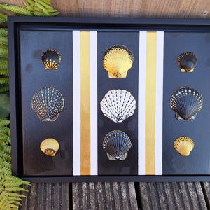 May include: A rectangular black framed wall art piece featuring nine decorative seashells arranged in a grid. The background consists of vertical black, white, and gold stripes. The shells are painted in black, white, and metallic gold patterns.