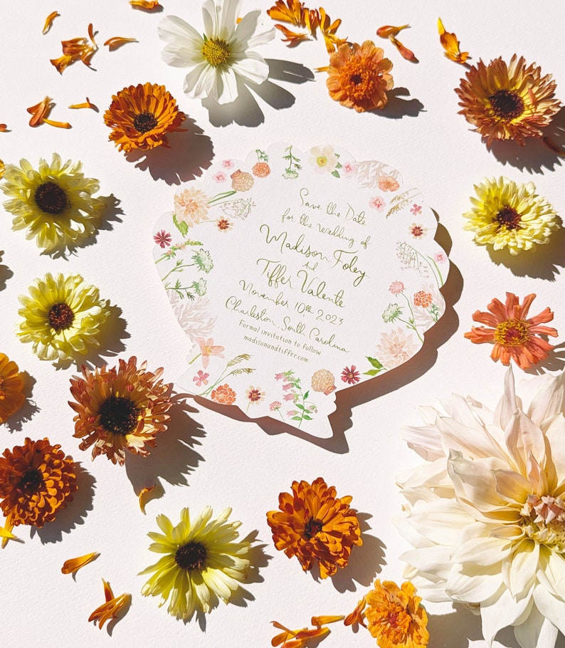 May include: Save the Date card featuring a floral design in the shape of a seashell. The card is for the wedding of Madison Foley and Tiffer Valente on 10th November 2023 in Charleston, South Carolina. The text "madisonandtiffer.com" is printed at the bottom of the card.
