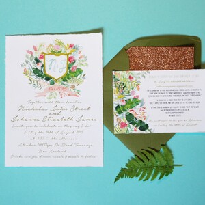May include: A wedding invitation with a tropical theme. The invitation features a watercolour illustration of a floral wreath with a shield in the centre. The shield has the initials "N.J." and the date "14.08.15". The invitation is printed on textured paper and has a pink ribbon.