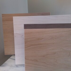 May include: Three wood samples, one with a dark brown border, stacked on top of each other. The top sample is a light brown wood, the middle sample is a white wood, and the bottom sample is a medium brown wood.