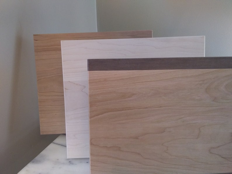 May include: Three wood samples, one with a dark brown border, stacked on top of each other. The top sample is a light brown wood, the middle sample is a white wood, and the bottom sample is a medium brown wood.