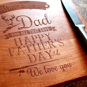 May include: A wooden cutting board with an engraved message that reads "Thank you Dad - For all that love - Happy Father's Day - We love you". The message is surrounded by a deer with antlers, a rifle, and a fishing rod.