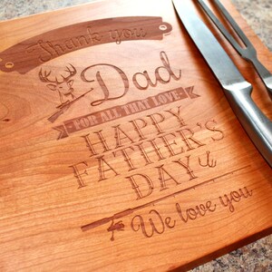 May include: A wooden cutting board with an engraved message that reads "Thank you Dad for all that love - Happy Father's Day - We love you". The board also features an engraved image of a deer with antlers and a rifle, and a fishing rod.