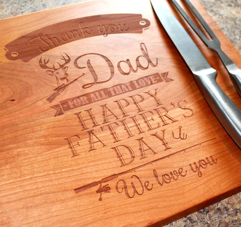 May include: A wooden cutting board with an engraved message that reads "Thank you Dad for all that love - Happy Father's Day - We love you". The board also features an engraved image of a deer with antlers and a rifle, and a fishing rod.