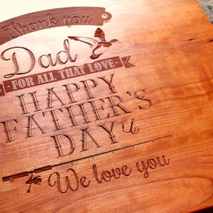 May include: A wooden cutting board with an engraved message that reads "Thank you Dad - For all that love - Happy Father's Day - We love you". The message is surrounded by a stylized fishing rod and a bird.