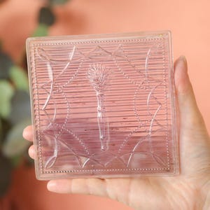 May include: A clear, square glass block with an etched floral design, held in a hand. The block features a central flower and linear patterns. The background is a soft peach color, with greenery visible on the left.