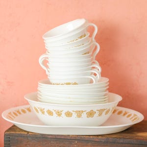 May include: A stack of white teacups with handles, saucers, and bowls with a gold floral design. The dishes are arranged on a white oval platter and a wooden surface, against a peach-colored wall. A vintage kitchenware set.