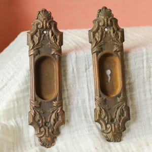 May include: Two antique bronze-colored pocket door pulls with ornate floral and scroll detailing. Each pull has a rectangular recessed area and a keyhole. The pulls are displayed on a white, textured fabric.