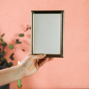 May include: A rectangular gold-colored picture frame with a white interior is held up against a peach-colored background. The frame has a decorative border. Green foliage and a candle are visible in the background.