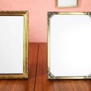May include: Two ornate, gold-colored picture frames with white interiors stand on a wooden surface. The frames have detailed, decorative edges. One frame is rectangular, and the other has rounded corners.