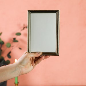 May include: A hand holds a gold picture frame with a white background. The frame has a decorative border.