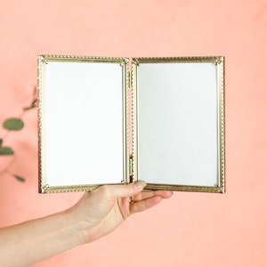 May include: A gold-colored picture frame with a beaded border is open to reveal two empty white spaces. The frame is being held in a hand against a light pink background.