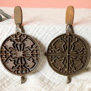 May include: Antique cast iron waffle iron with wooden handles. The round iron is open, revealing the intricate patterned interior. The waffle iron is brown and sits on a white cloth.