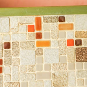 May include: A rectangular surface featuring a mosaic pattern of small, varied tiles in shades of beige, tan, and cream, interspersed with vibrant orange, deep red, and dark brown square and rectangular accents, finished with a solid olive green border.